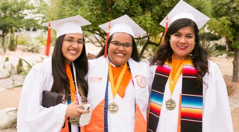 Three female graduates in Scott Courtyard, two with Chicano/Latino stole and one with 卡帕 stole. Karen Kandamby is in the center; others are unidentified.